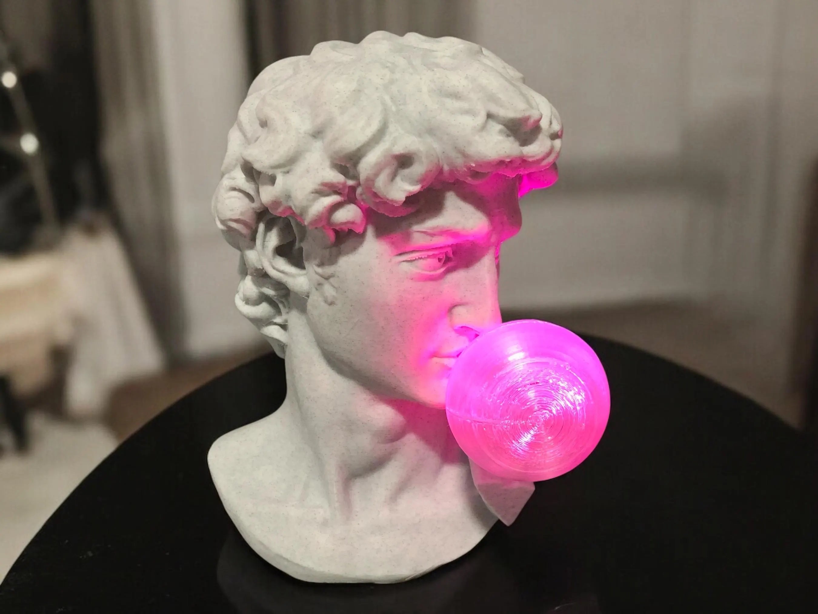 Marble bust of a classical figure blowing a pink bubble with a blurred indoor background